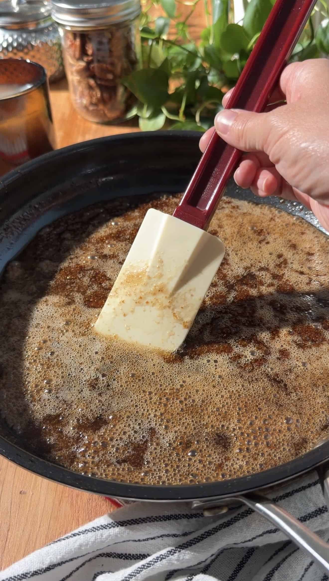A hand stirring brown sugar and butter in a saucepan with a silicone spatula.