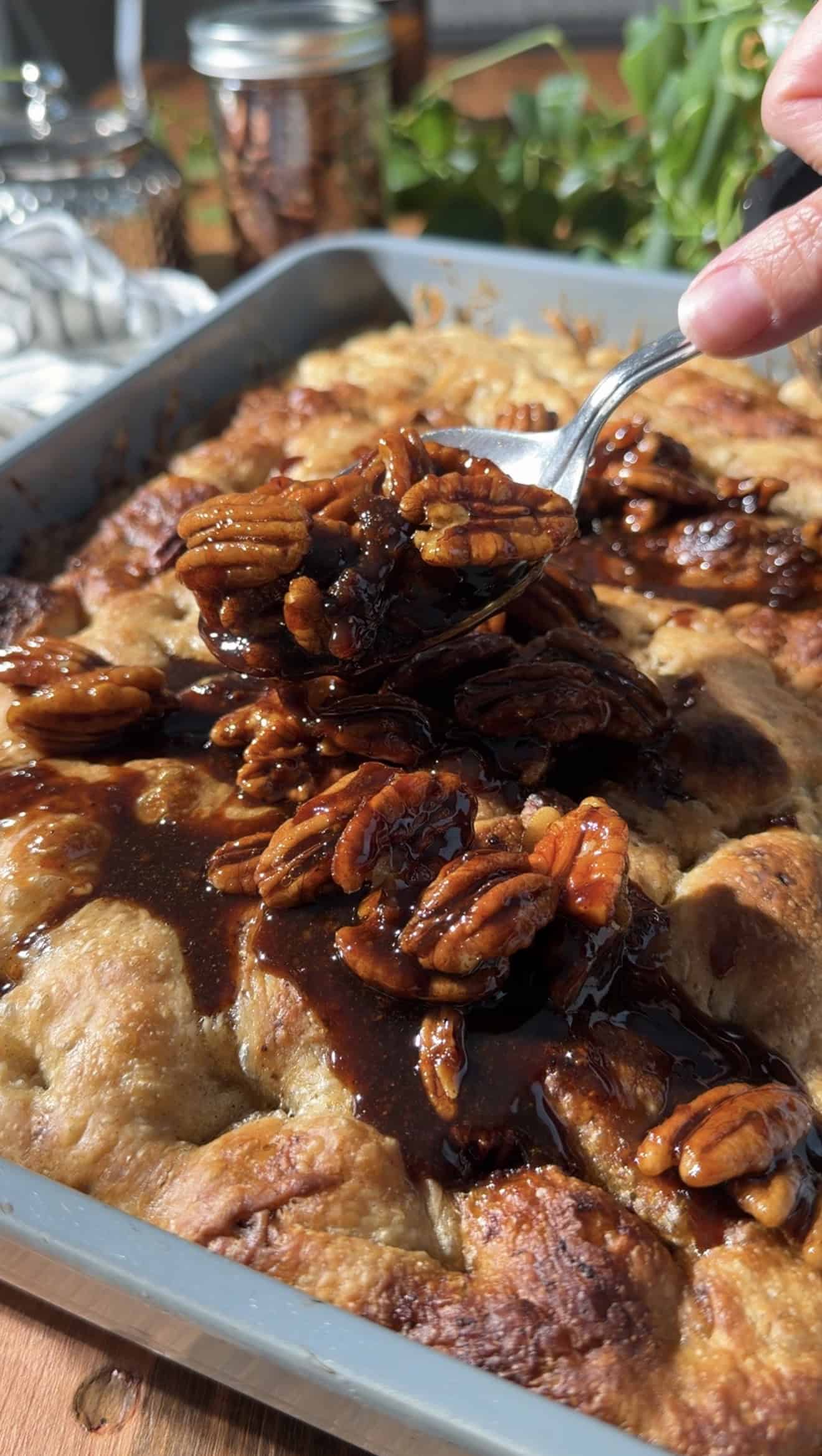 A hand spooning candied whole pecans in brown buttery sauce onto baked focaccia in a metal dish.