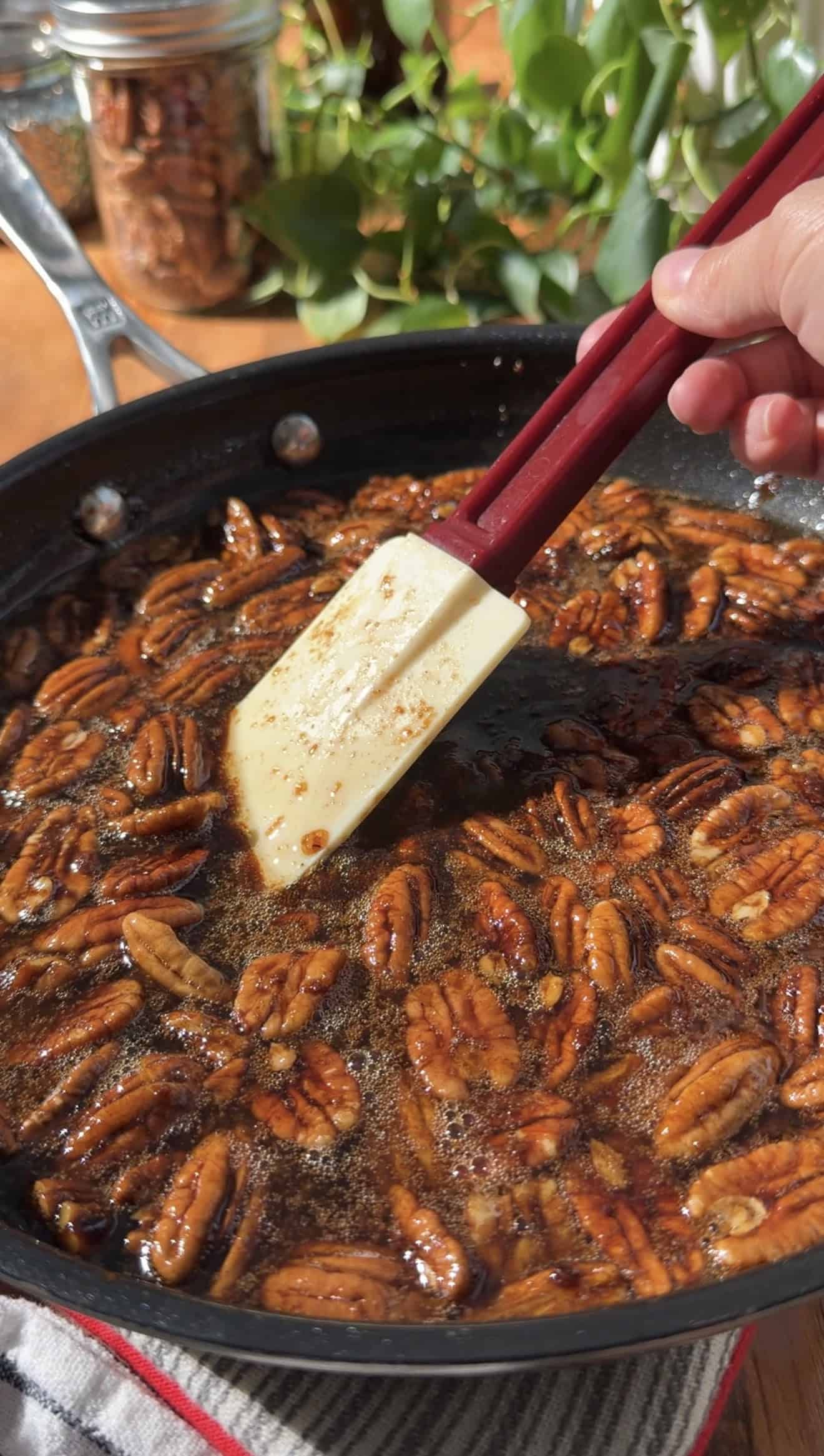 A hand stirring candied whole pecans in a brown sugar butter sauce in a saucepan.